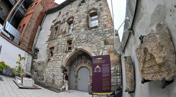 Ein Mann betrachtet die mittelalterliche Steinfassade eines Gebäudes, das Teil des Weltkulturerbes Erfurt ist. Daneben sind alte Steintafeln mit hebräischen Inschriften ausgestellt.