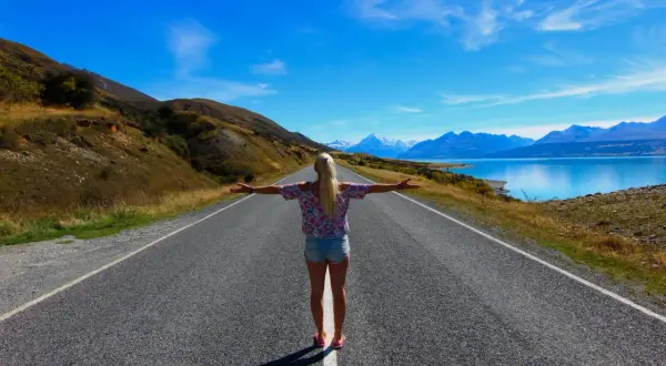 Frau mit ausgebreiteten Armen auf einer Straße, die zu schneebedeckten Bergen und einem blauen See führt. Ein Traum von Work and Travel Weltreise.