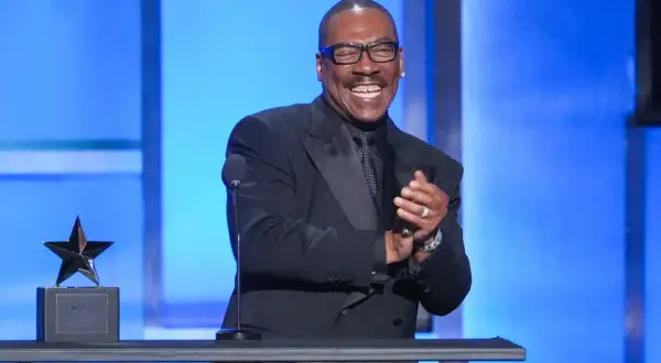Eddie Murphy, känd för sina roliga filmer, ler och klappar händerna på scenen bredvid en stjärnstatyett.