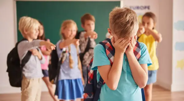 Smutny chłopiec zakrywa twarz, podczas gdy inne dzieci wskazują na niego palcami. To obraz tego, co zrobić, gdy dziecko jest wyśmiewane w szkole.