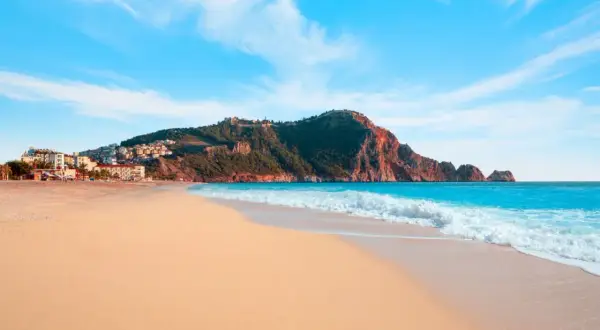 Plaża Kleopatry w Alanyi, z turkusowym morzem, złotym piaskiem i malowniczymi wzgórzami z zamkiem.