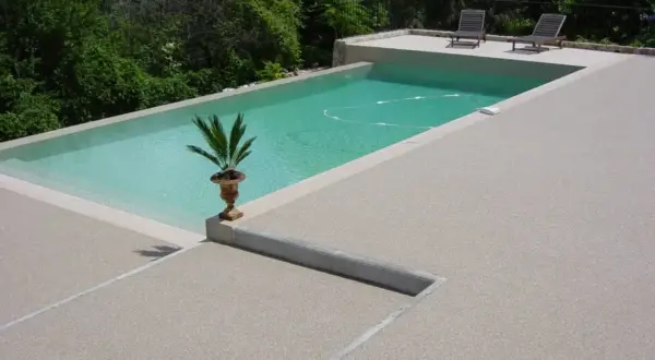 Piscina con bordo in cemento e pavimentazioni in cemento a vista, circondata da vegetazione lussureggiante.