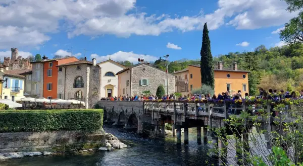 Un pittoresco borghetto mantovano con case colorate, un ponte di legno affollato di gente e un fiume che scorre sotto un cielo azzurro con nuvole bianche.