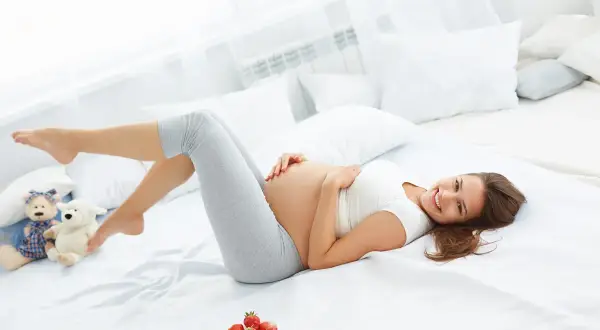 Uśmiechnięta ciężarna kobieta leży na łóżku, głaszcząc brzuch. Obok pluszowe misie i miseczka truskawek. Czasem dokucza jej zespół niespokojnych nóg w ciąży.