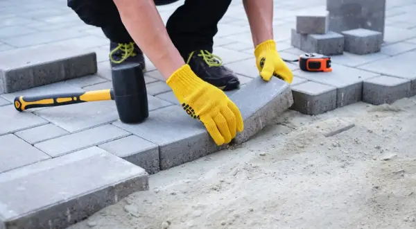 Układanie kostki brukowej na podjazd. Ręce w żółtych rękawiczkach precyzyjnie dopasowują kolejny element. Jaka grubość kostki na podjazd będzie najlepsza?