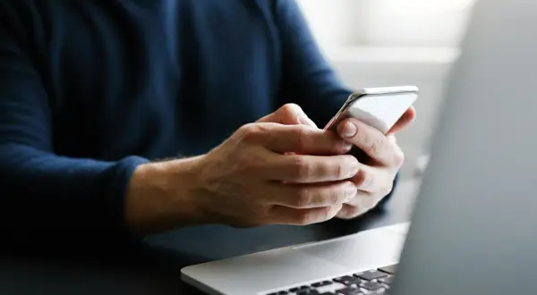Mężczyzna trzyma telefon, szukając informacji, jak przenieść kontakty na kartę SIM. Obok leży laptop.