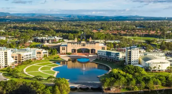 Weite Luftaufnahme des Campus der Bond University mit einem zentralen See, gepflegten Grünflächen und modernen Gebäuden, umgeben von Bäumen und Hügeln.