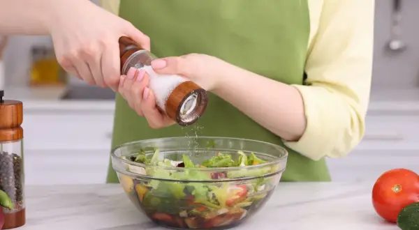 Manos sazonando una ensalada con sal, uno de los usos de la sal en la cocina.