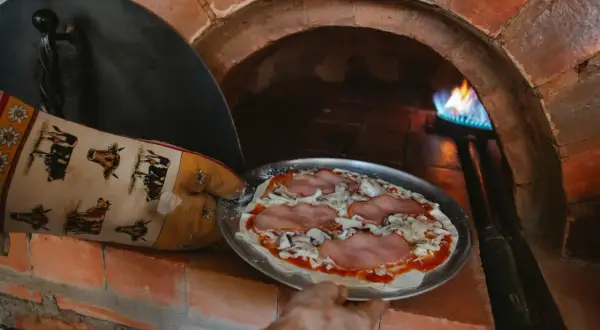 Ręka w rękawicy z krowami wkłada pizzę do pieca szamotowego. Płomień tańczy w głębi pieca.
