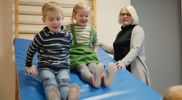 Zwei Kinder rutschen auf einer blauen Matte, während eine Erzieherin zusieht. Ein Fall könnte eine aufsichtspflichtverletzung kita darstellen.