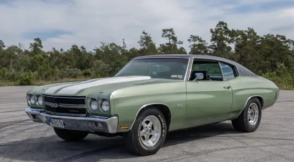 Grön Chevelle SS från 1970 med vita ränder på huven, parkerad utomhus med träd i bakgrunden.
