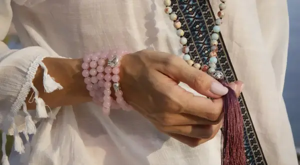 Mano con varios tipos de japa mala: uno de cuarzo rosa en la muñeca y otro largo con cuentas de colores y borla.