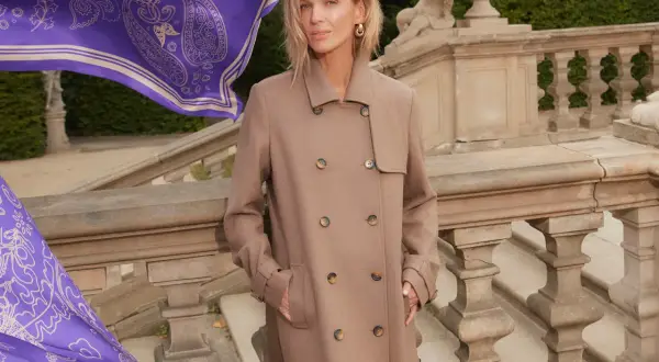 Elegancka kobieta w beżowym płaszczu, obok niej powiewa fioletowy szal z wzorem. Zastanawiasz się, jaki kolor pasuje do fioletu? Beż jest doskonałym wyborem!