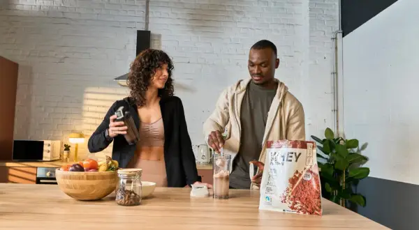 Ein Mann und eine Frau bereiten einen Proteinshake zu. Die Packung zeigt, wie wird Proteinpulver hergestellt.