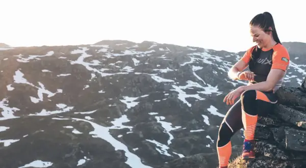 Une femme en tenue de sport s'assoit sur des rochers, regardant sa montre, prête à trouver un trail dans un paysage montagneux enneigé.