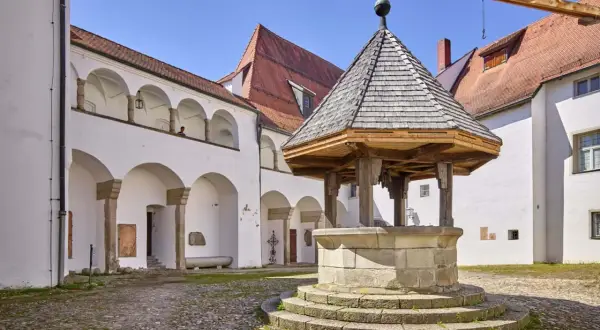 Historischer Brunnen im Innenhof des Oberhaus Passau. Weiße Arkadenbögen und rote Ziegeldächer unter blauem Himmel.