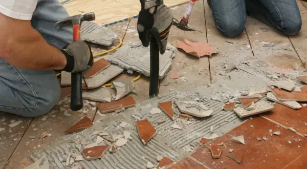 Deux personnes s'y prennent pour comment décoller du carrelage au sol, utilisant marteaux et burins pour briser les carreaux et le ciment.