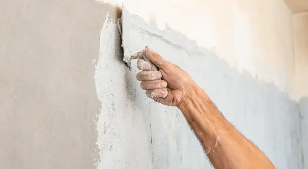 Mano che applica intonaco spugnato su una parete con una spatola. La texture è grezza e irregolare.