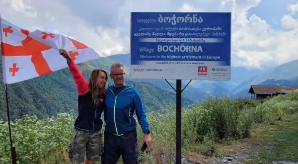 Para podróżników z Gruzji, pytanie "czy w Gruzji jest bezpiecznie" nabiera nowego znaczenia. Para z gruzińską flagą na tle górskiego krajobrazu i tablicy informującej o najwyżej położonej wsi w Europie.