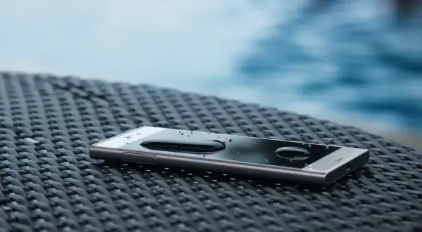Telefon z kroplami wody na ekranie, leżący na plecionej powierzchni przy basenie. Klasa szczelności **IP 65** zapewnia ochronę przed zachlapaniem.