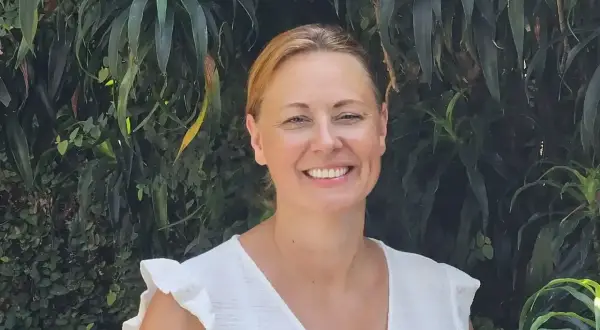 Vrolijke vrouw met blonde haren, lachend in een witte jurk, tegen een achtergrond van weelderig groen. Arjan van Dam zou trots zijn op deze foto.