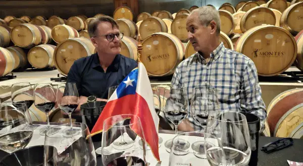 Dwóch mężczyzn przy degustacji wina w piwnicy pełnej beczek. Flaga Chile i kieliszki sugerują, że to degustacja win Montes.