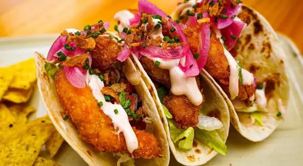 Tres deliciosos tacos de pescado crujientes, cubiertos con cebolla morada encurtida, salsa cremosa y cilantro fresco.