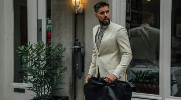 Mann im cremefarbenen Anzug mit gemustertem Hemd und Krawatte, trägt eine schwarze Kleidersacktasche. Ein lässiges Outfit für unterwegs.