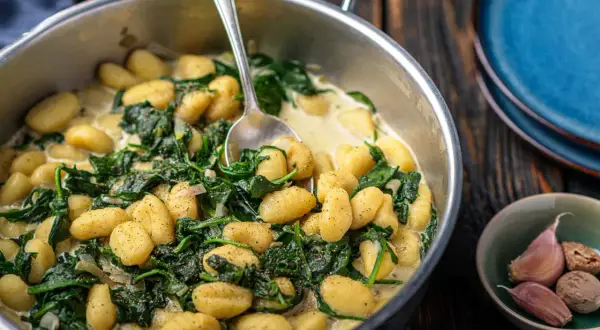 Gnocchi ze szpinakiem w kremowym sosie, podane w metalowej patelni, obok czosnku i gałki muszkatołowej.