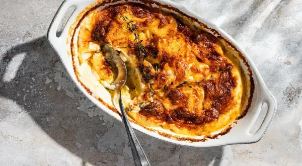 Ein goldbraun gebackenes Kartoffelgratin in einer weißen Auflaufform, garniert mit frischem Thymian. Eine silberne Löffelspitze lugt aus der cremigen Füllung hervor.