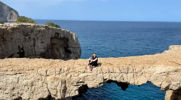 Una mujer sentada en un arco de roca natural, el "cala columpio ibiza", con el mar azul de fondo.