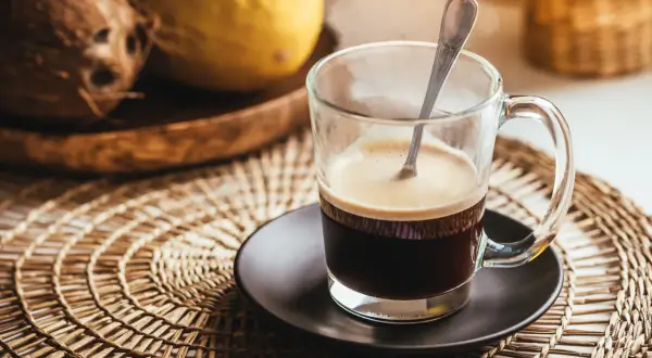 Tazza di caffè fumante su sottobicchiere intrecciato. Sullo sfondo, cocco e melone. Un caffè lungo ha più caffeina per iniziare la giornata.