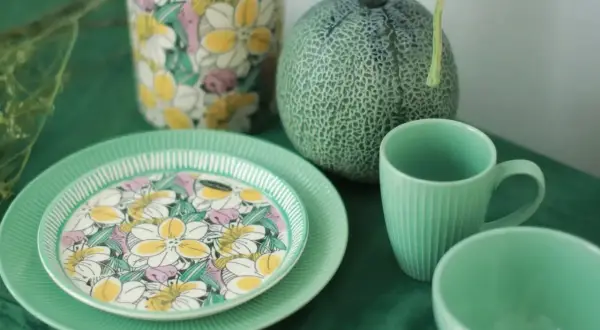 Assiette en bone china porcelaine ornée de fleurs colorées, à côté d'une tasse et d'un bol verts, avec un melon et un vase fleuri.