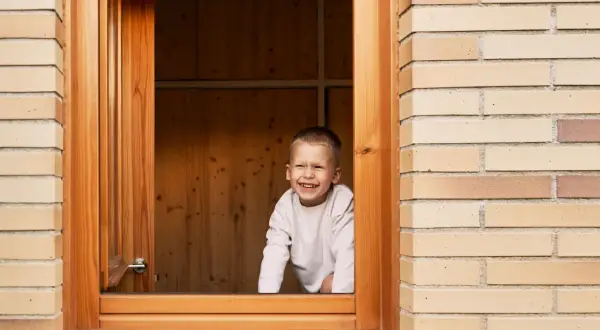 Niño sonriente asoma por una ventana de madera recién instalada.