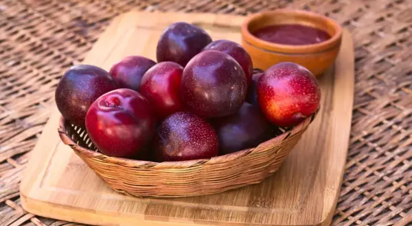 Korb mit dunklen, glänzenden Pflaumen. Vorsicht: unreife Pflaumen giftig! Daneben eine kleine Schale mit Pflaumenmus.