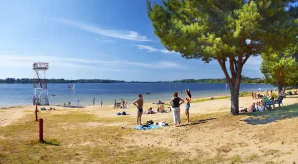 Słoneczna plaża nad jeziorem goczałkowickim. Ludzie odpoczywają, kąpią się i pływają na żaglówkach. Widoczna wieża ratownicza.