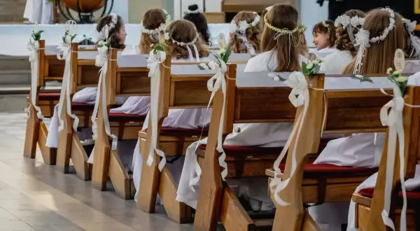 Dzieci w bieli, z wiankami na głowach, siedzą w ławkach kościelnych. Czekają na uroczystość, zastanawiając się, ile trwa komunia.