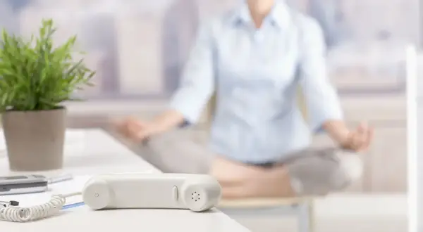 En person mediterar på kontoret. En telefonlur och en växt syns i förgrunden. En pausgympa på jobbet kan ge ny energi.