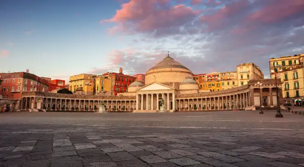 Piazza del Plebiscito a Napoli, con il Palazzo Reale e la Basilica di San Francesco di Paola, sotto un cielo rosato al tramonto.