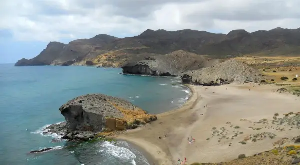 Paisaje de las mejores calas de Cabo de Gata, con formaciones rocosas, arena dorada y mar turquesa bajo un cielo nublado.