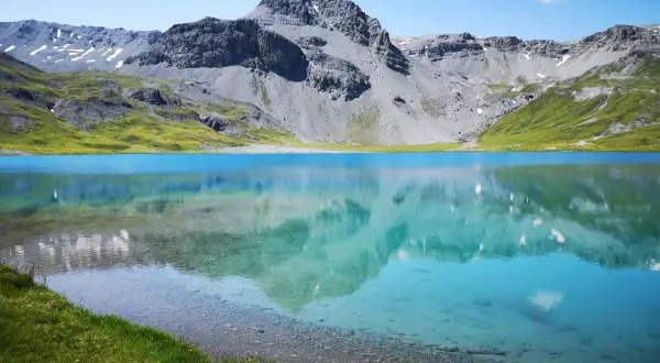 Ein klarer, blauer Bergsee, der **lai da rims**, spiegelt die majestätischen, grauen Berge und grünen Hänge wider.