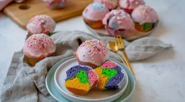 Bunte regenbogen muffins mit rosa Zuckerguss und Streuseln, ein Stück auf einem Teller zeigt das farbenfrohe Innere.