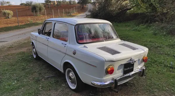 Vit Simca-bil parkerad på en gräsbevuxen stig, med olivträd och åkermark i bakgrunden.