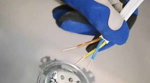 Ręka w rękawicy trzyma przewody elektryczne, przygotowując się do podłączenia fazy w gniazdku. Widoczne są przewody brązowy, niebieski i żółto-zielony.