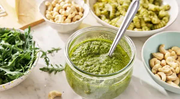 Świeże pesto z rukoli w słoiku, obok miseczki z orzechami nerkowca, rukolą i kawałkiem parmezanu.