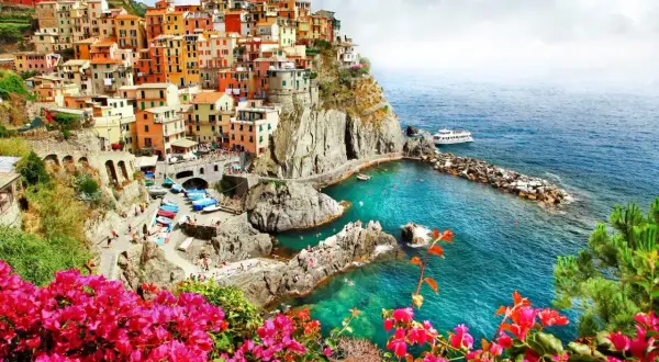 Färgstarka hus klättrar uppför klipporna vid den italienska kusten. Klart turkost vatten och rosa blommor ramar in vyn.