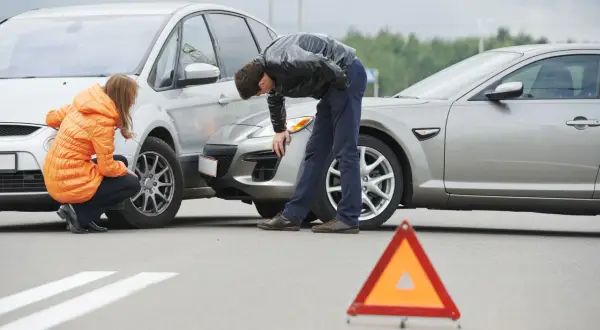 Incidente stradale con due auto danneggiate. Una donna in giacca arancione e un uomo esaminano i veicoli. Triangolo di segnalazione a terra. Nessun segno di omissione di soccorso.