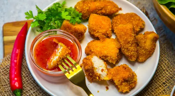 Knusprige Hähnchen-Nuggets, die man einfach selber machen kann, mit süß-saurer Soße und frischer Petersilie. Ein roter Chili liegt daneben.