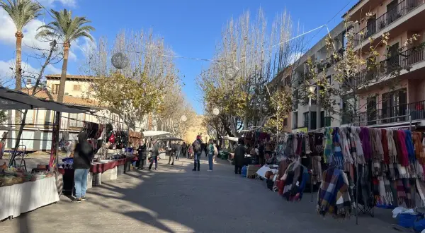 Livlig marknad i Alcudia med stånd som säljer kläder och accessoarer. Palmträd och byggnader kantas av gatan.