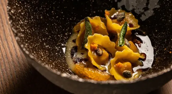 Tortelli di zucca dorati, guarniti con salvia croccante e arancia, in una ciotola scura e rustica.